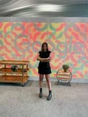 Kamilly, wearing a black dress and shoes, stands with her arms crossed in front of a multi-colored wall with the Google logo. To her right is a multi-tiered wooden table with a plant on the middle shelf, and to her left is a multi-tiered shelf with a plant on top.