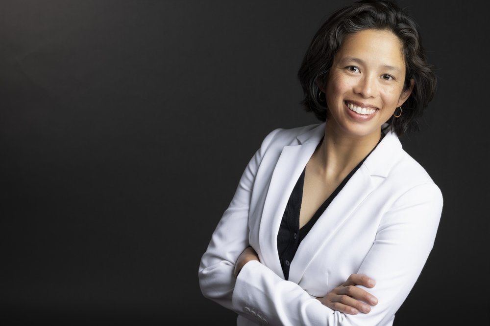 This is a photo of a woman in a white jacket in front of a black background with her arms crossed.