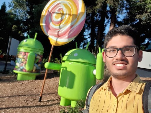 Kaustubh, wearing a yellow shirt and glasses, stands outside in front of two green Android statues. One statue is filled with candy, the other is holding a lollipop and holding up a peace sign.