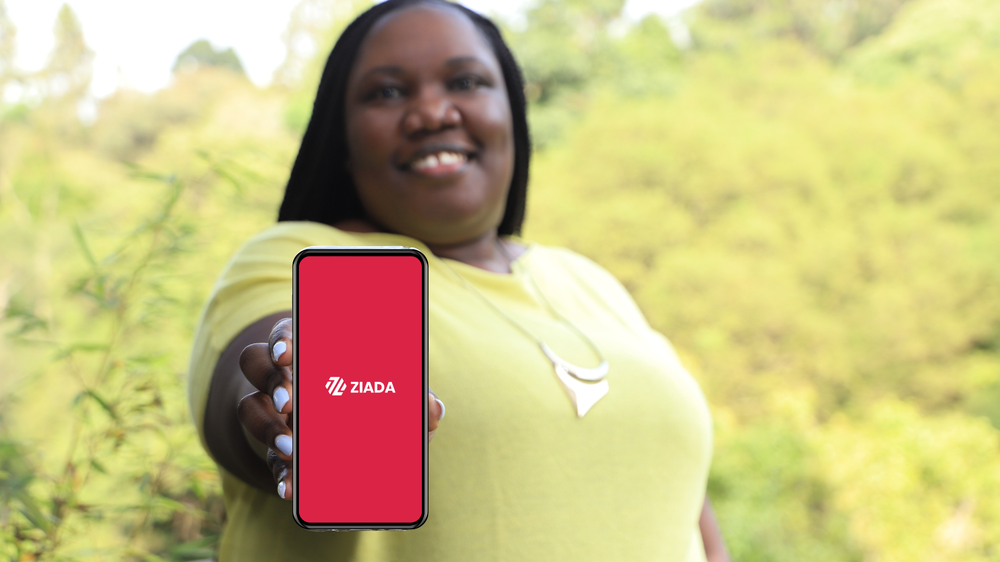 A person wearing a yellow short-sleeved shirt smiles and holds a phone showing the Ziada logo on the screen.