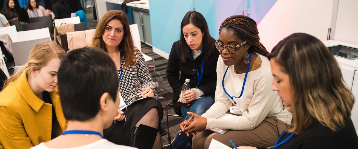 A group of six women founders sitting on chairs in a circle, talking, with other founders and more chais visible in the background.