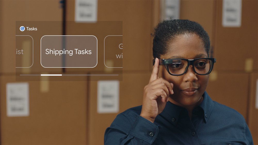 A woman with black hair touches her Google Glass Enterprise Edition eyewear while standing behind stacked cardboard boxes. She selects the Tasks function on her device, with an image popping up in her vision.