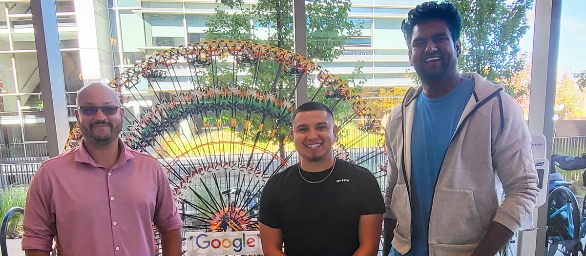 Three men stand in front of a model Ferris wheel that says “Google” in a brightly-lit office. Out the large window behind them you can see grass, trees, and a window-paneled building.