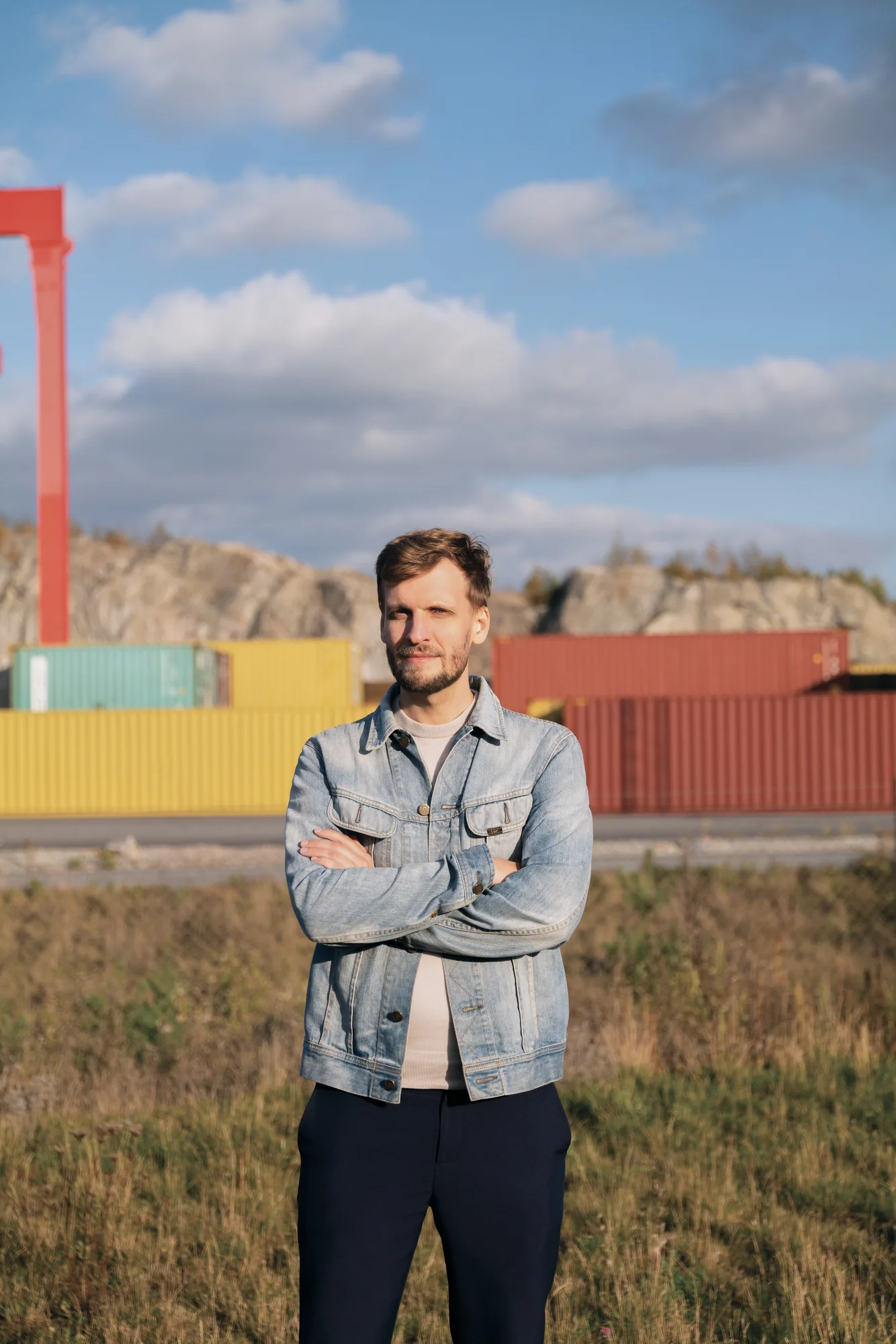 Headshot of Kristian wearing a jean jacket and standing in front of a moving cargo train with red, yellow and blue shipping containers.