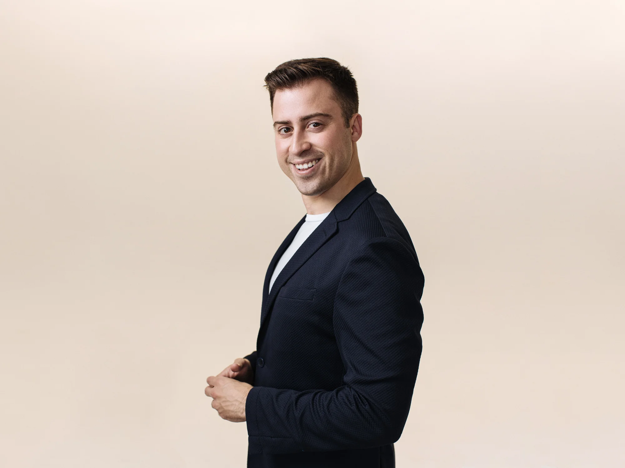 A man with short dark hair wearing a black suit jacket and white shirt smiles at the camera, in front of a gray background.