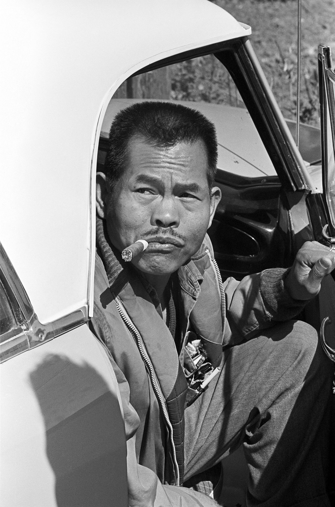 A black-and-white photograph of a man sitting in the passenger seat of a car. The car door is open; he looks back with his left hand on the car door and a cigar in his mouth. In the foreground, a shadow is visible on the car.