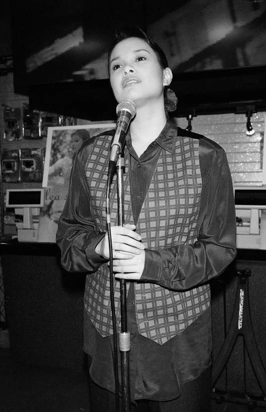 A black-and-white photograph of a woman, looking upwards while singing into a standing microphone. She is wearing a long-sleeved shirt with a patterned vest.