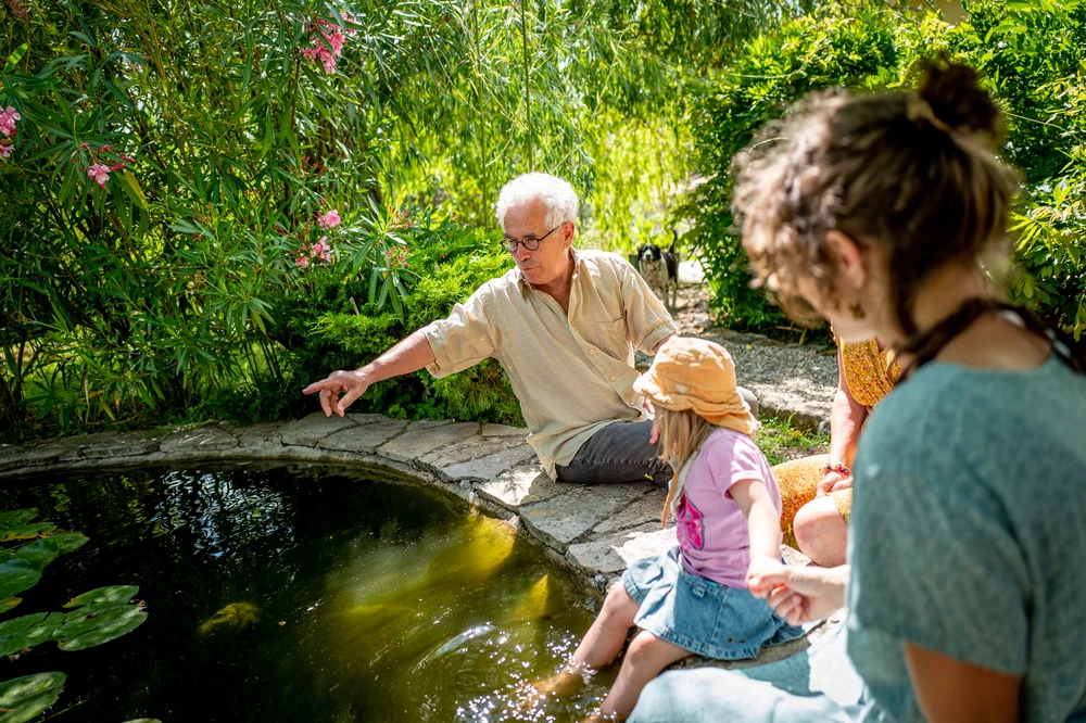 A man sits with a woman and a young child at French cottage Les Courtines, pointing towards a pond.