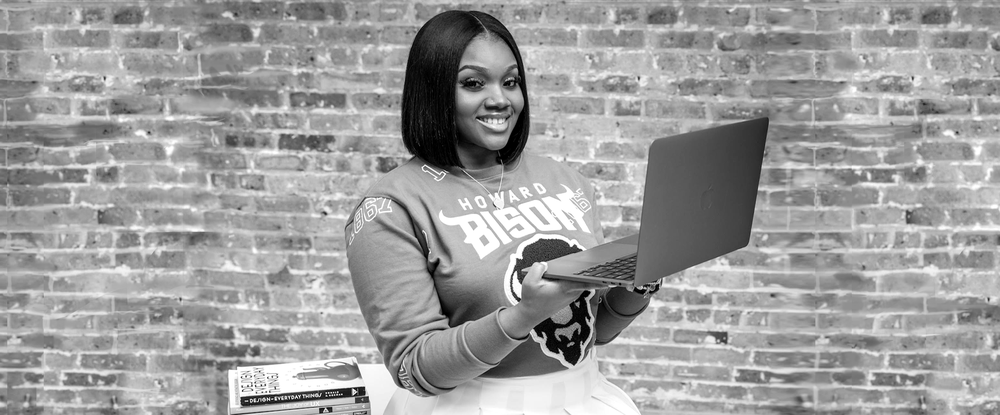 A black-and-white photo of Leslie Coney posing in front of a stack of books, wearing a Howard University sweater and holding a laptop.