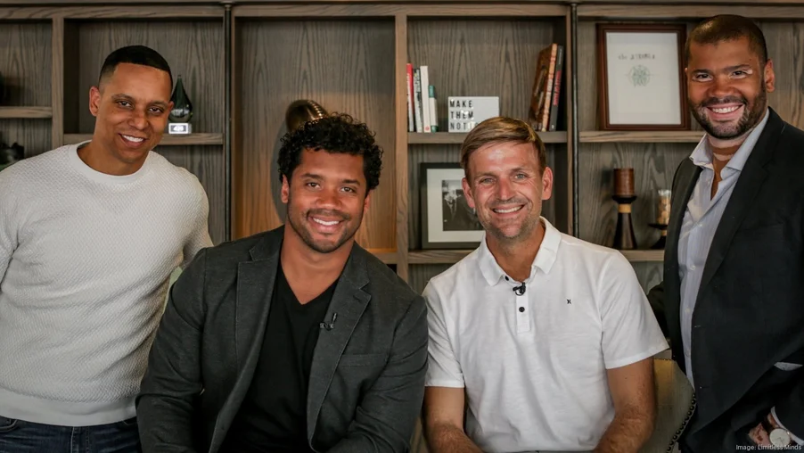 Four men— two wearing white t-shirts and two wearing jackets— look at the camera as they stand in front of a bookshelf.