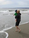 An older brother holds a younger brother while walking on the beach.