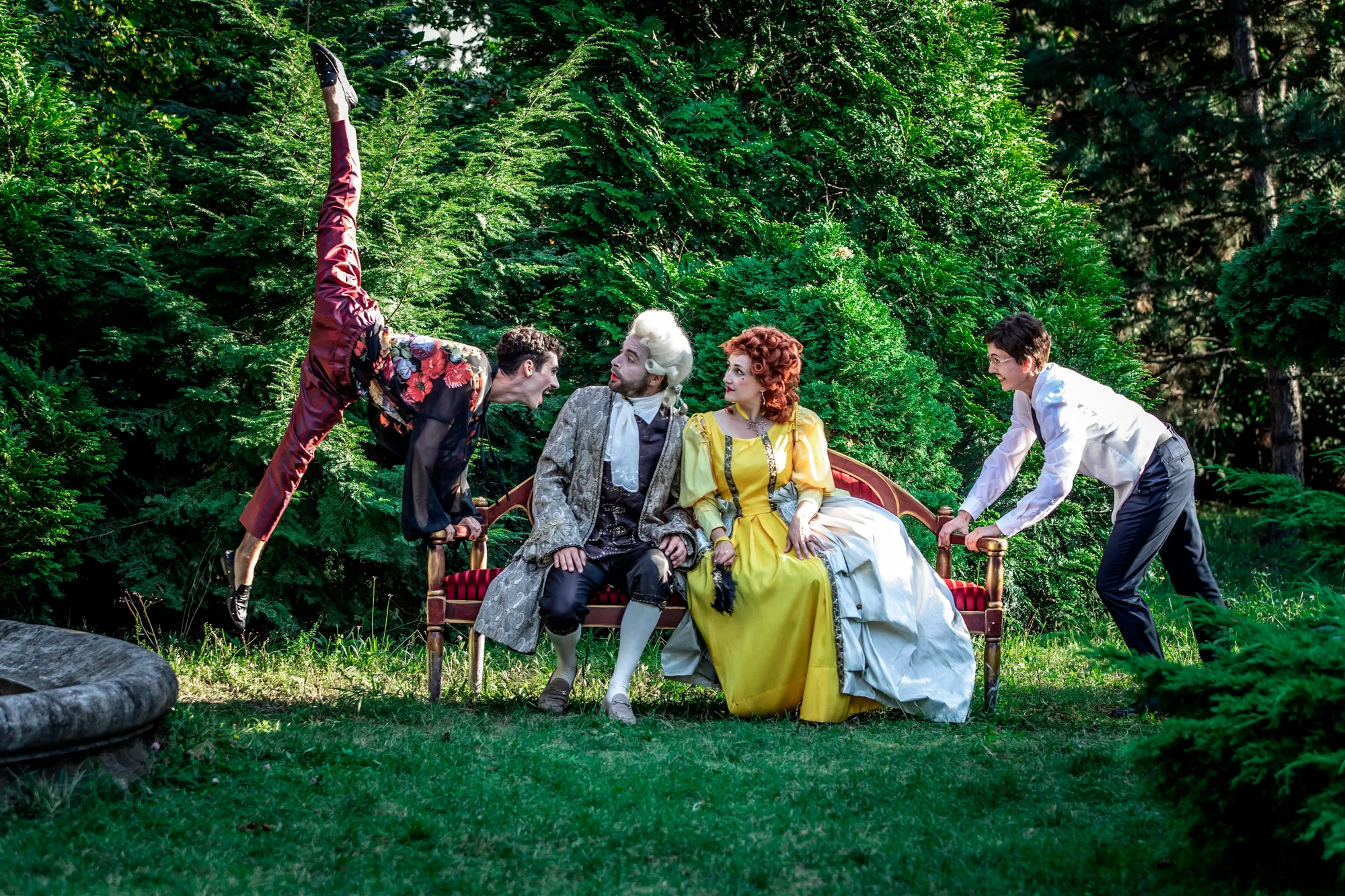 The picture depicts a theatrical scene set in a lush, outdoor environment. Four individuals are engaged in a dramatic pose. A person on the left is performing an impressive, acrobatic move with one leg extended high in the air. This individual is dressed in colorful, ornate clothing. Two individuals are seated on a red, antique-style bench. The man in the center wears 18th-century style clothing. The woman next to him wears a vibrant yellow dress with period-specific details, including a voluminous skirt and intricate design. On the right, a person in a modern white shirt and dark pants is bent forward, holding the arm of the bench, appearing to be in motion or exerting effort.