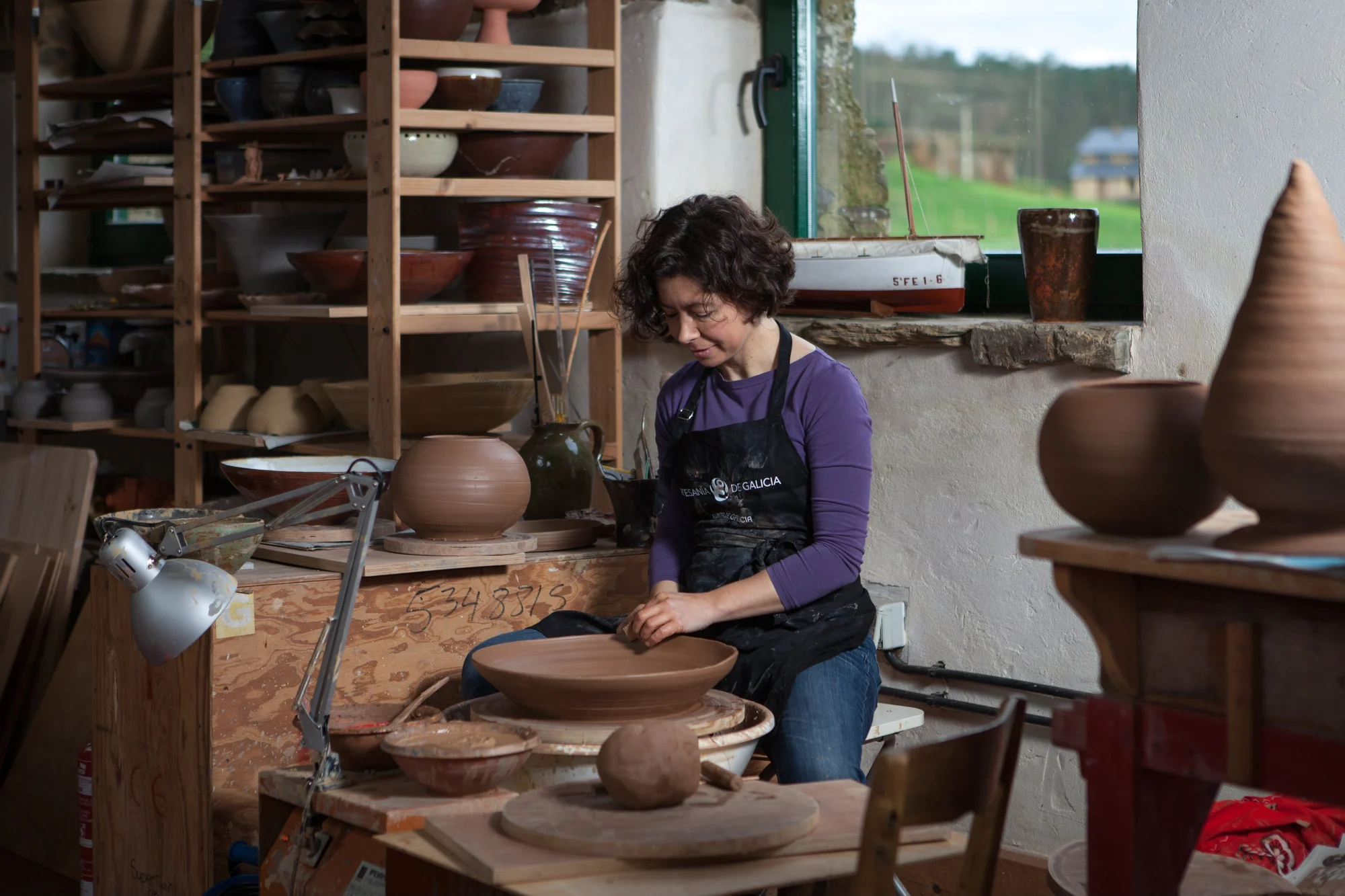 Photograph of a woman working on a pottery wheel, surrounded by ceramic pieces. There is a window in the background.