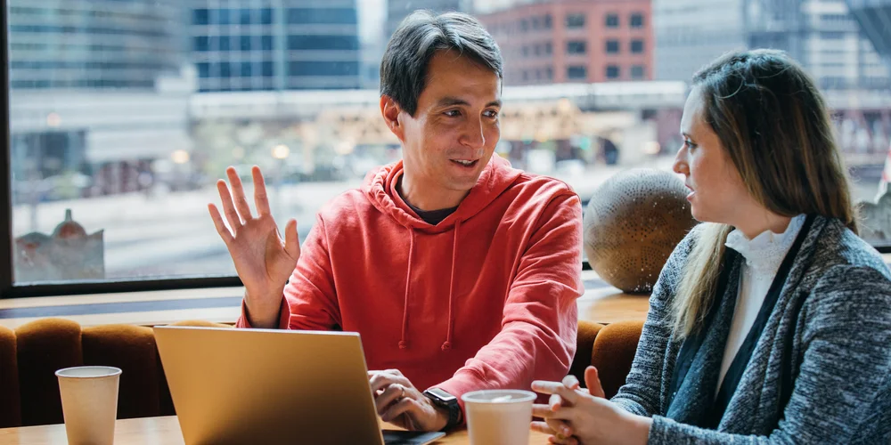 Founder Luis Suarez, wearing a red sweatshirt, sits at a table talking with Google mentor Victoria Bujny. They have a laptop and coffee cups in front of them and the Chicago skyline is visible out the window behind them.