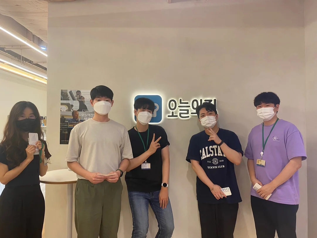 A group of five people in masks lean against a wall and look at the camera