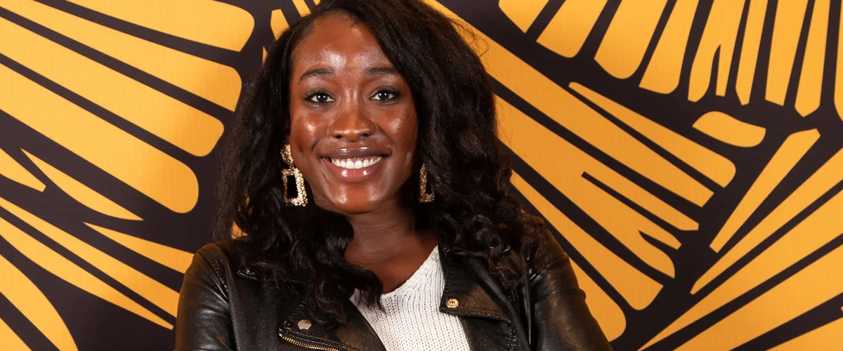 Ada, with crossed arms and wearing a black leather jacket, smiles at the camera against a yellow and black backdrop