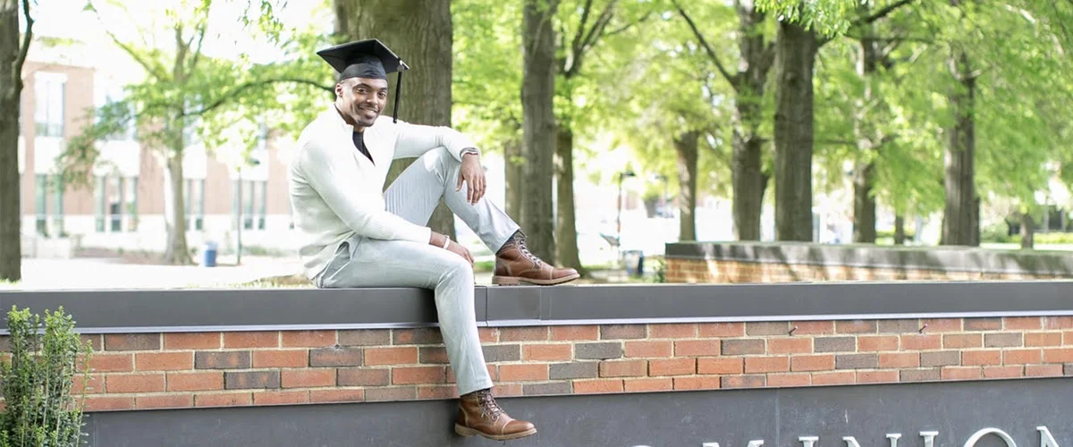 Dre’ wears a graduation cap while sitting on a wall, with trees and buildings in the background.