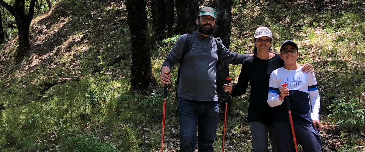 Manav standing with his wife and son while out hiking in a wooded area. They are all carrying red walking sticks.