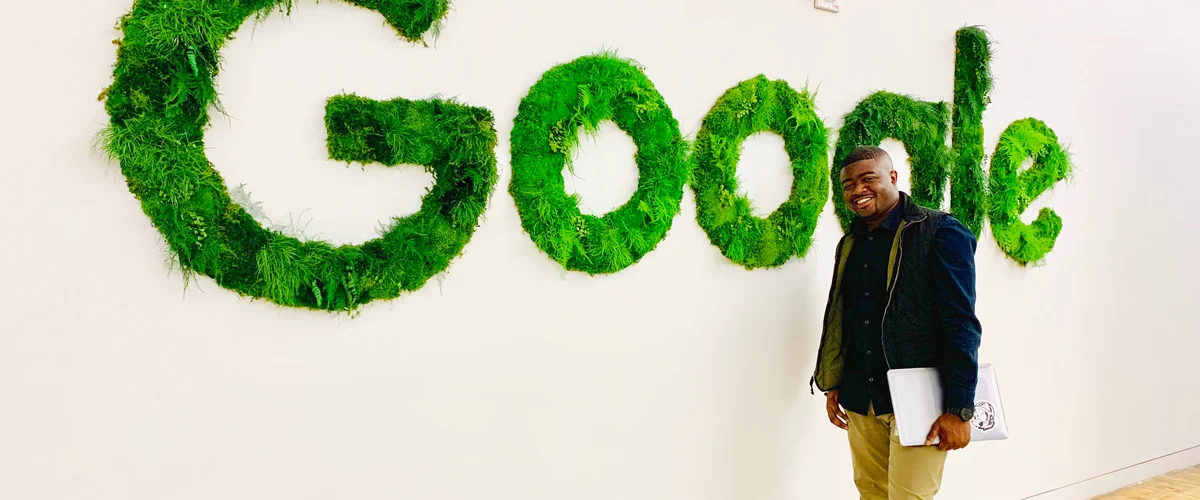 Rob holding a laptop and standing next to white wall with large sign made of grass and moss that spells out the word, “Google.”