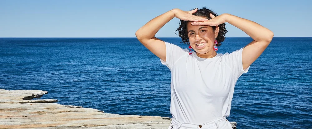 Tamina standing near the edge of a flat rock surface with the ocean behind her. She is wearing a white top, white pants and red and blue earrings. She is using her hands to shield her eyes from the sun.