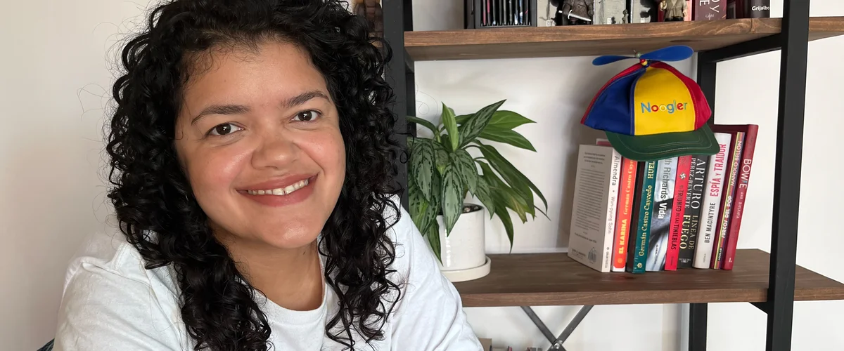 Paula smiling in her home office. Behind her is a bookcase with a Noogler hat, plant and books.