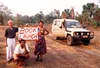 Image of three people standing in the desert holding a sign that says “Book Launch' next to an old four wheel drive car