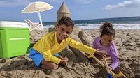 Photo of two young kids building a sand castle on the beach. In the background there is a chair and umbrella and bright green cooler. The chair and umbrella are selected using Magic Eraser.