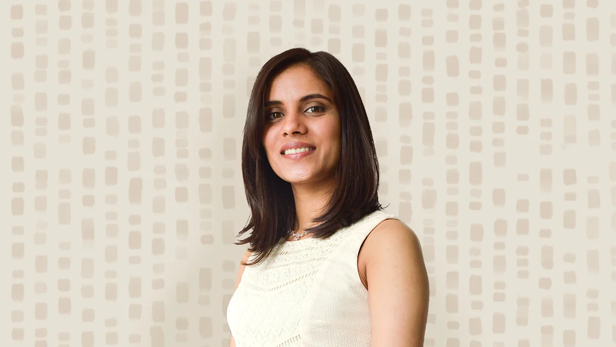 A woman with shoulder-length brown hair and a white shirt stands in front of a beige illustrated background