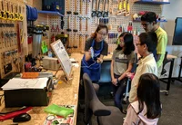 A Googler shows four children how to use a tool attached to a cell phone. In the background are construction tools like hammers, mallets, pliers and saws. In the foreground is a sketch of a cartoon character, a mouse and laptop.