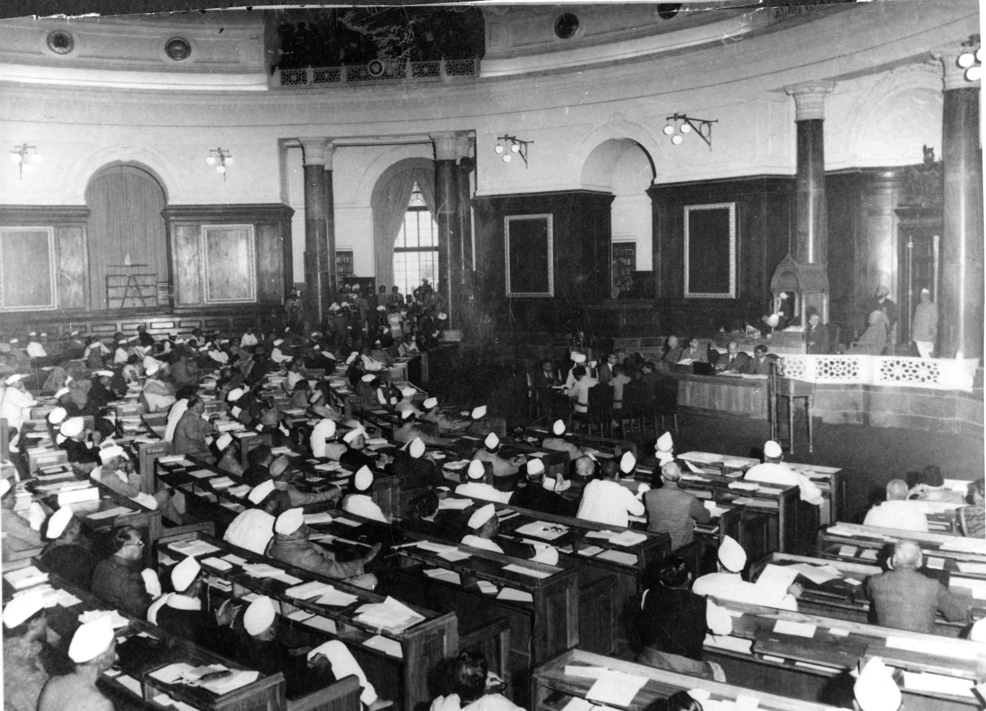 Black and white photo of a circular room with windows, with rows of people sitting on benches and looking at loose papers.