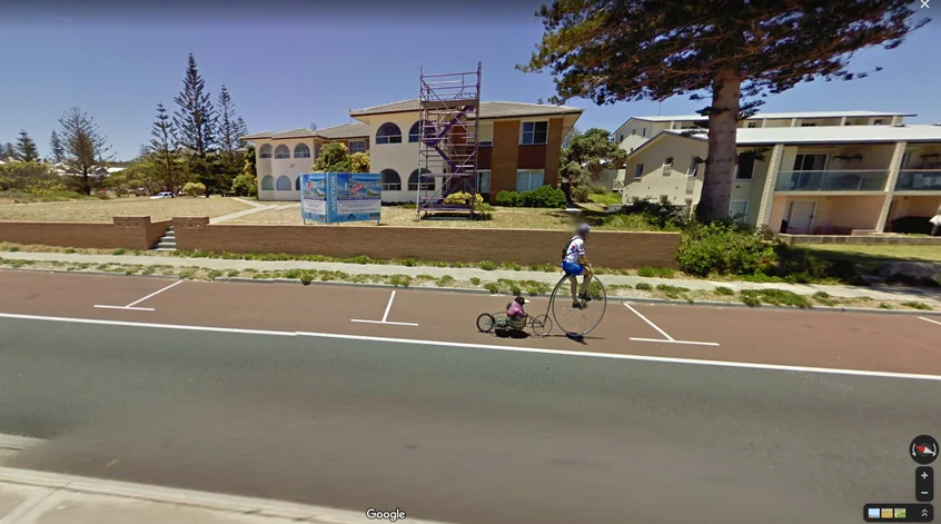Street View imagery showing a man riding on a high wheel bike in Cottesloe, WA – with a toy penguin riding at the back.