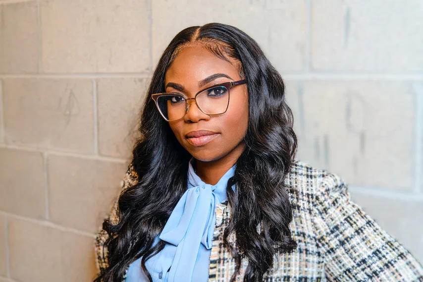 A Black woman with long dark hair and wearing glasses looks at the camera, with a light blue top and a brown plaid blazer.