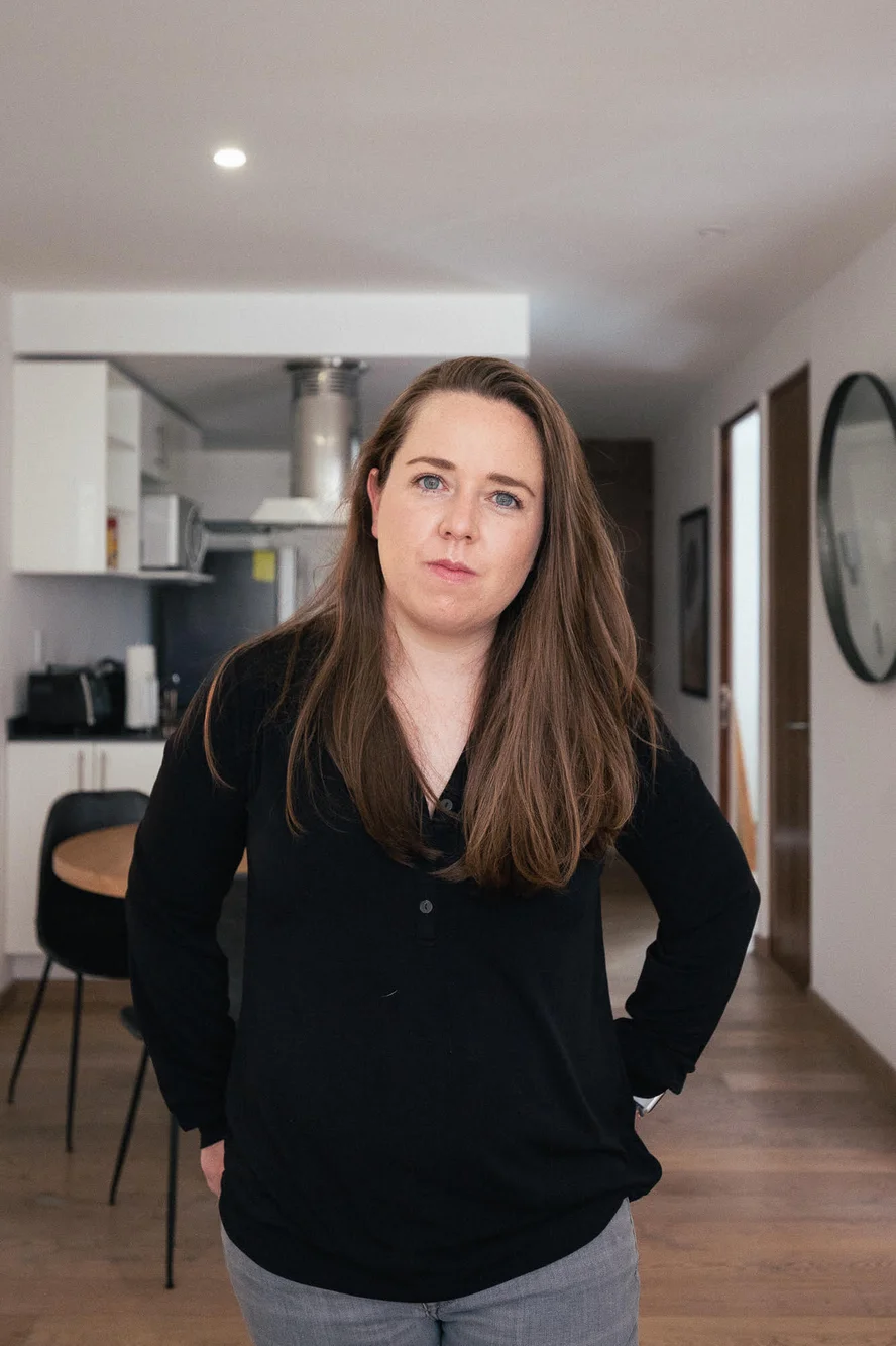 A woman standing in front of the camera wearing a black shirt. In the background there is a kitchen and a mirror, both are out of focus.