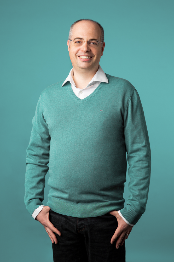 A man wearing glasses, a teal sweater and black pants stands in front of a teal background.