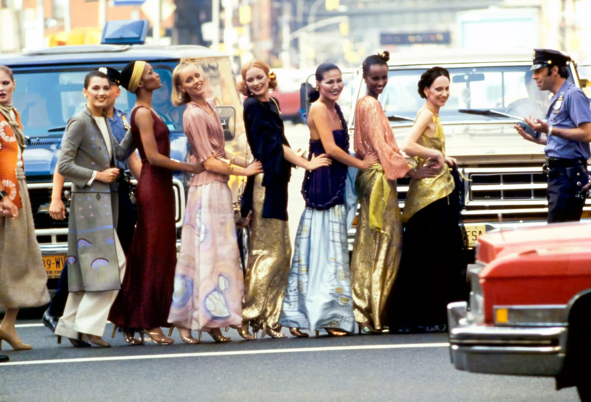 A group of models looks at the camera while crossing the street and holding on to one another, all wearing long colorful dresses and skirts