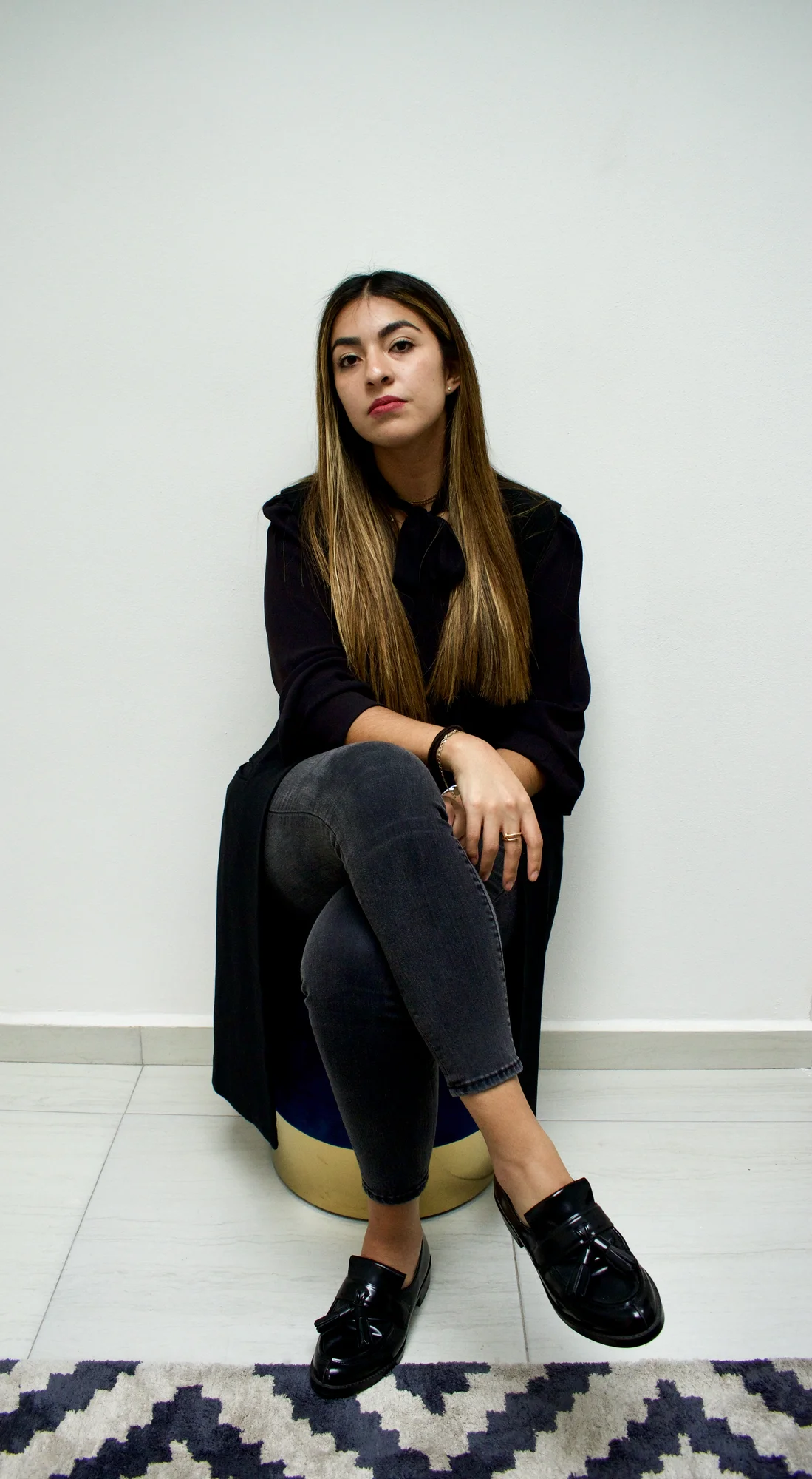 A woman dressed in black is sitting on a stool with her legs crossed. The background shows white walls and white tiled flooring, showing part of a zig-zag patterned rug.