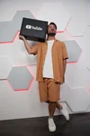 Manuel in piedi indossa un completo con short arancio e tiene sulla spalla una cassa con logo YouTube