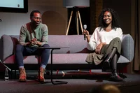 Clifford Ondara und Deborah Choi sitzen auf einer Bühne in Berlin auf einem grauen Sofa. Deborah hält ein Mikrofon in ihrer rechten Hand und lächelt.