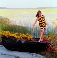 A model wearing a short yellow and black dress stands in a boat filled with flowers at the edge of a pond