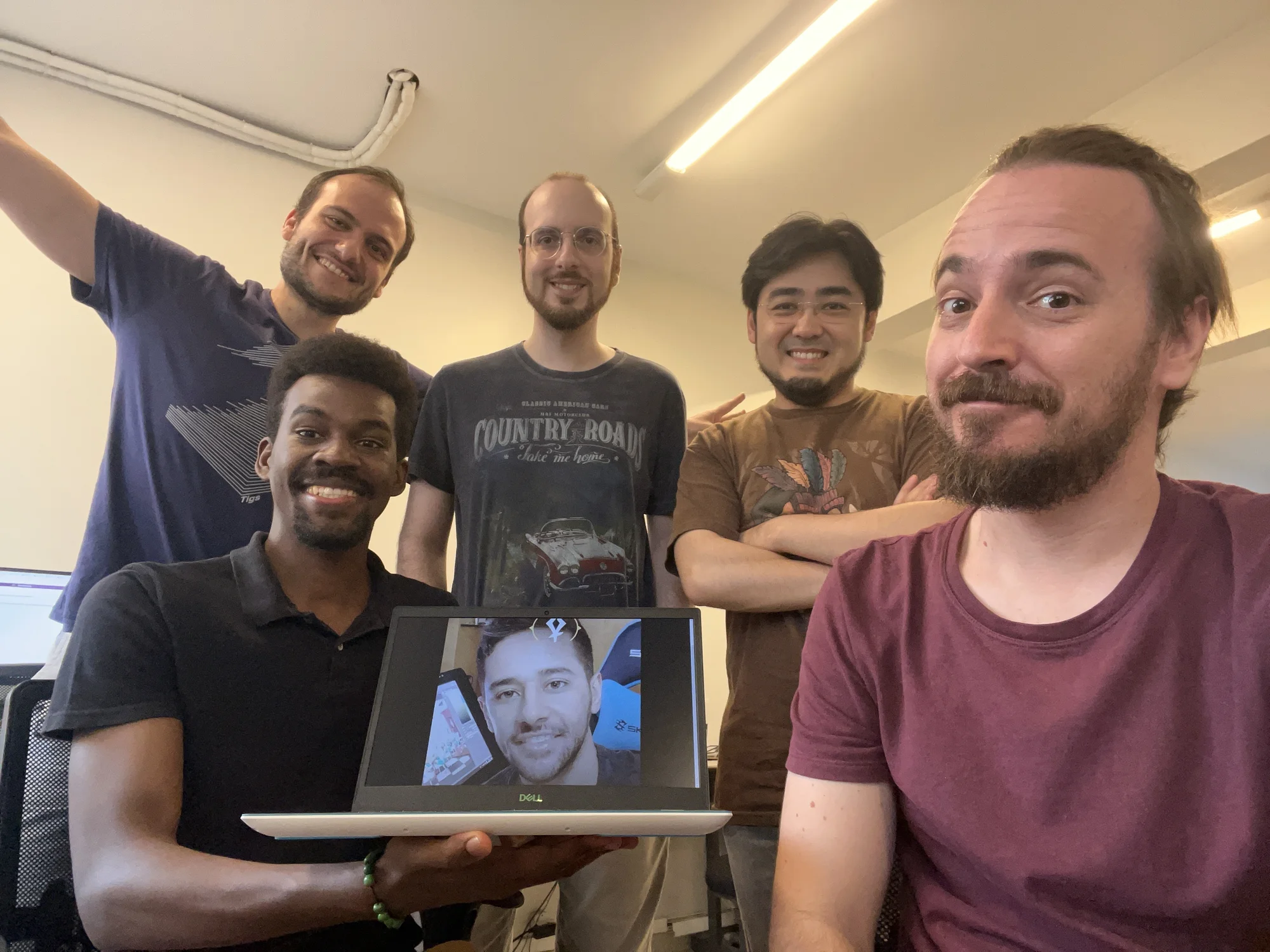 Five people stand around a laptop that shows a picture of another person’s face.
