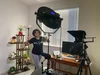 Priyanka in her work-from-home studio, which includes a lighting and camera setup, desk and display shelf of potted plants.