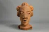 Image depicts a stylised sculpture of a female head. This figure has triangular eyes with holes in its eyes and mouth and hair parted into mounds. The sculpture is created in a light brown material with flecks of white .