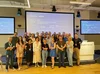 A group of about 40 people pose for a photo on a stage together wearing event lanyards and badges with projected screens behind them