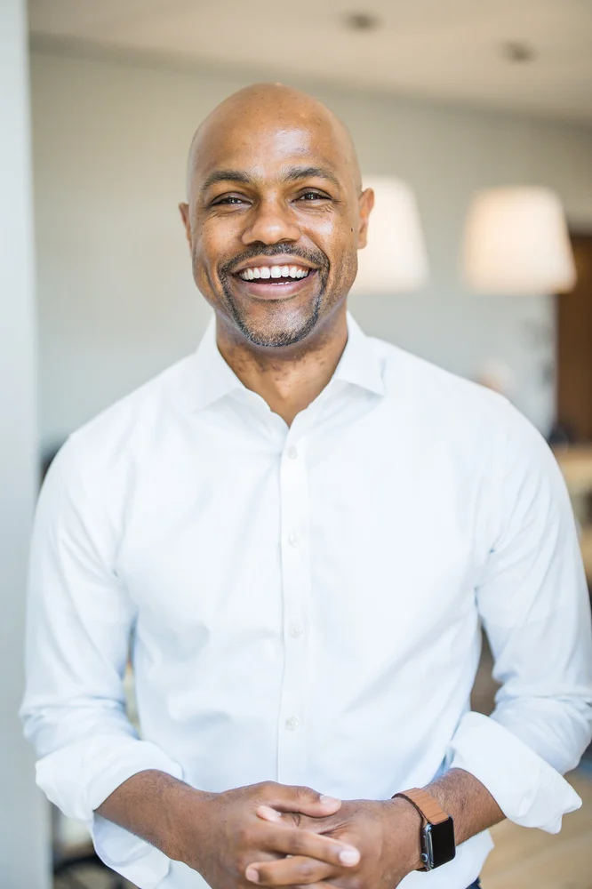 Nasir Qadree  smiles at the camera. He is wearing a white button down shirt. There are two lamps and a white wall behind him.