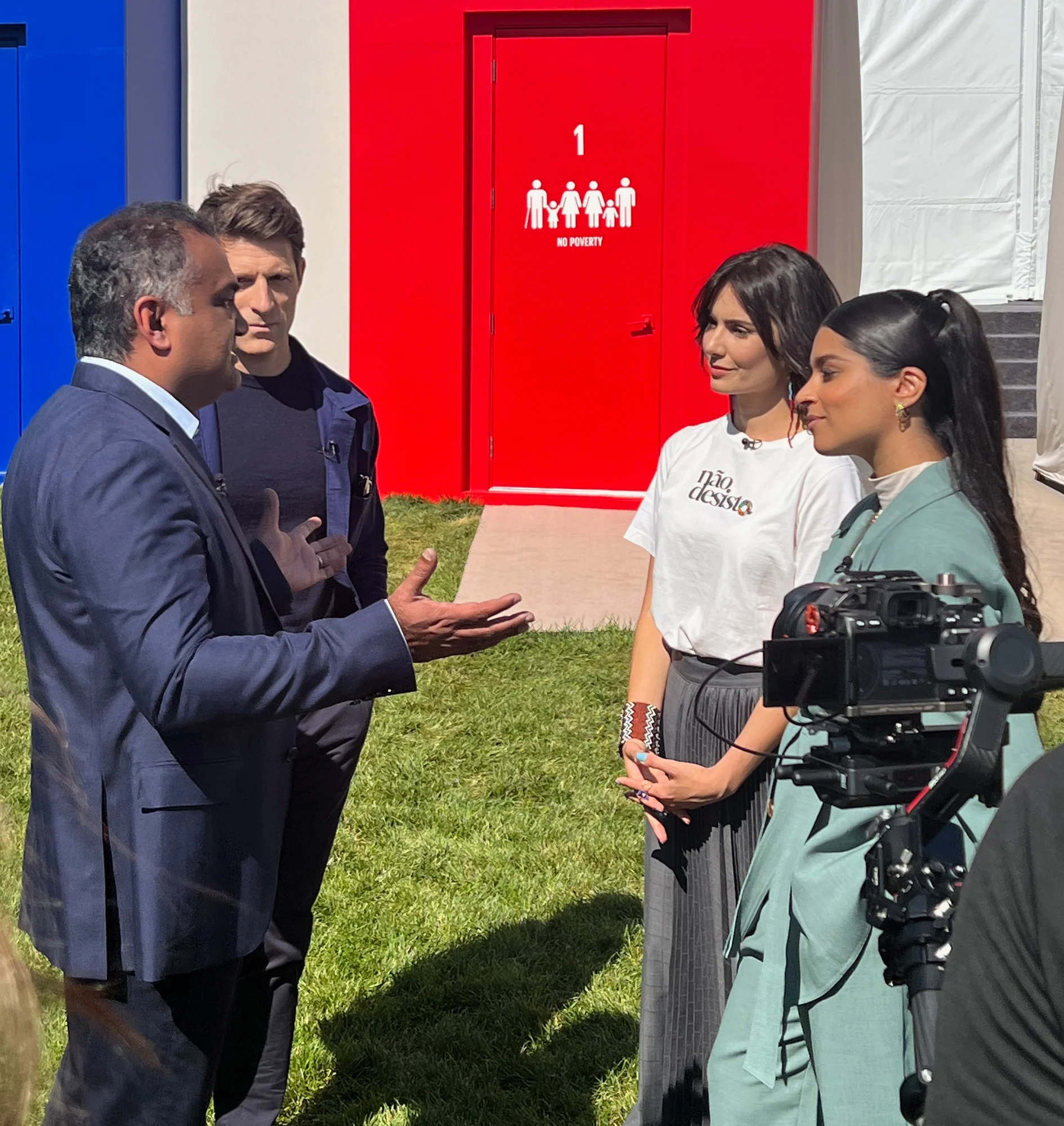 four people are having a conversation outdoors in front of a red door that reads "No poverty"