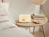 A picture of the Nest Hub with an orange morning glow, sitting on a night stand next to a bed.