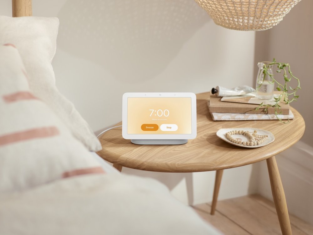 A picture of the Nest Hub with an orange morning glow, sitting on a night stand next to a bed.