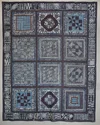 This image shows a  work comprised of sections of different fabrics which have been joined together to form a patchwork wall hanging. Indigo is the predominant color of this work, but white black and light blue are also present Each piece of cloth has geometric shapes woven into it.