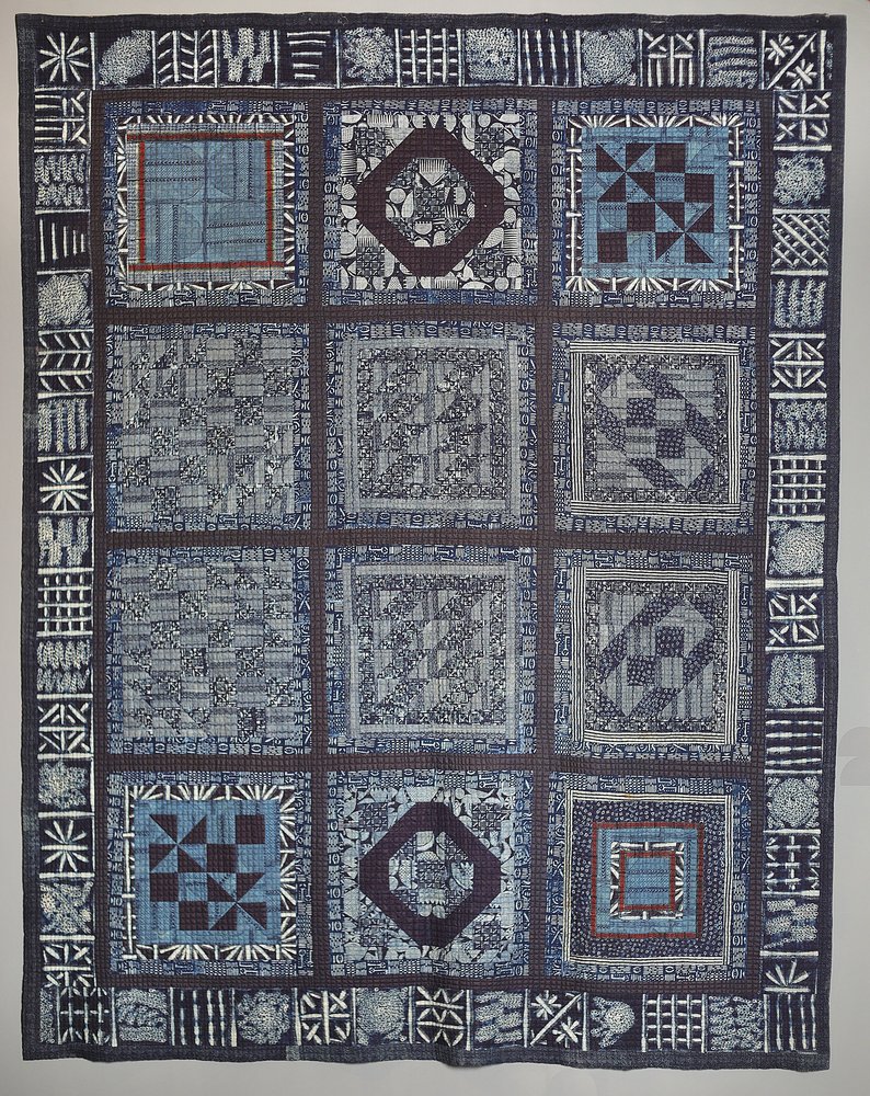 This image shows a  work comprised of sections of different fabrics which have been joined together to form a patchwork wall hanging. Indigo is the predominant color of this work, but white black and light blue are also present Each piece of cloth has geometric shapes woven into it.