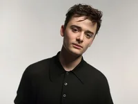 Photograph of Noah Schnapp looking directly to camera wearing a black shirt. Noah has white skin and brown short hair.